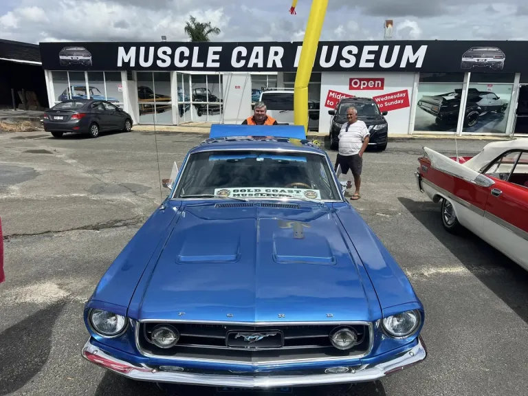 Blue Ford Mustang fastback with a Gold Coast Mustang club sign on the dash, parked outside the museum