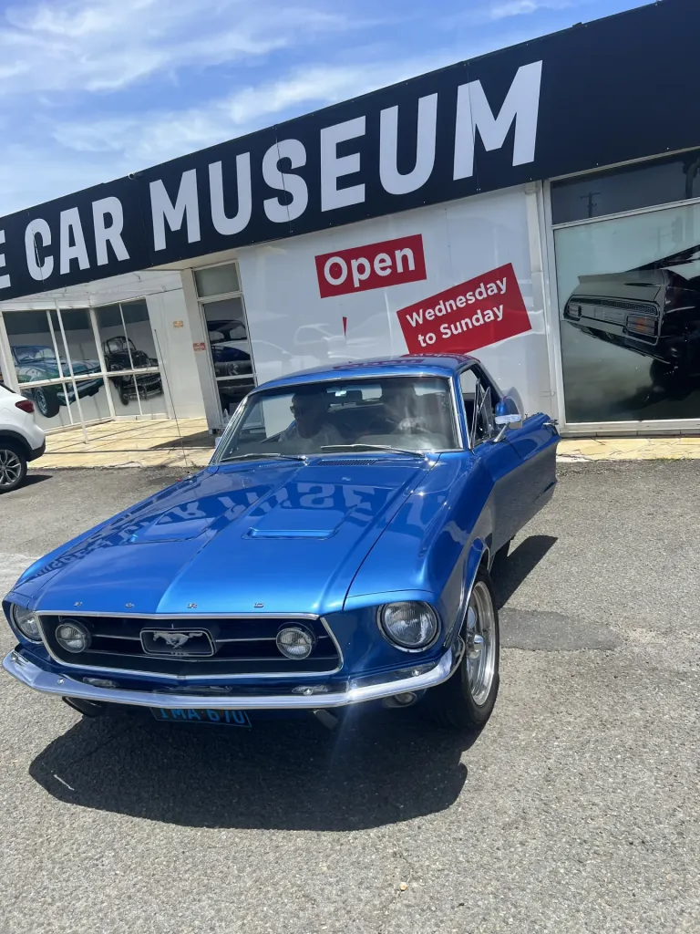 Blue classic Ford Mustang parked outside the Muscle Car Museum entrance under an Open sign