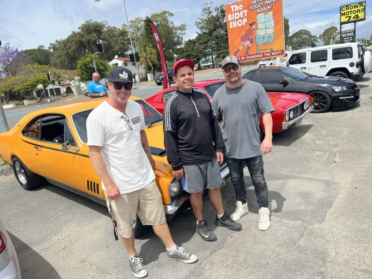 Three men posing with an orange classic coupe, a red classic coupe parked behind them
