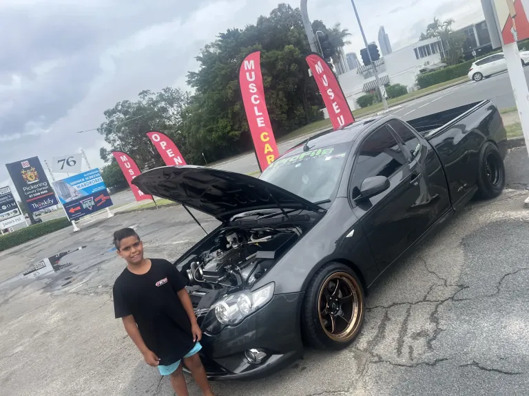 Boy standing beside a black ute with its bonnet open and bronze wheels near Muscle Car flag banners