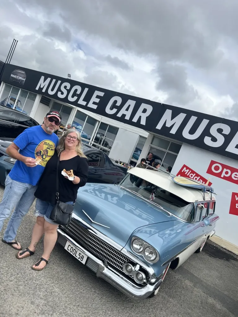 Couple holding food beside a pale blue 1950s station wagon with a surfboard on the roof outside the museum