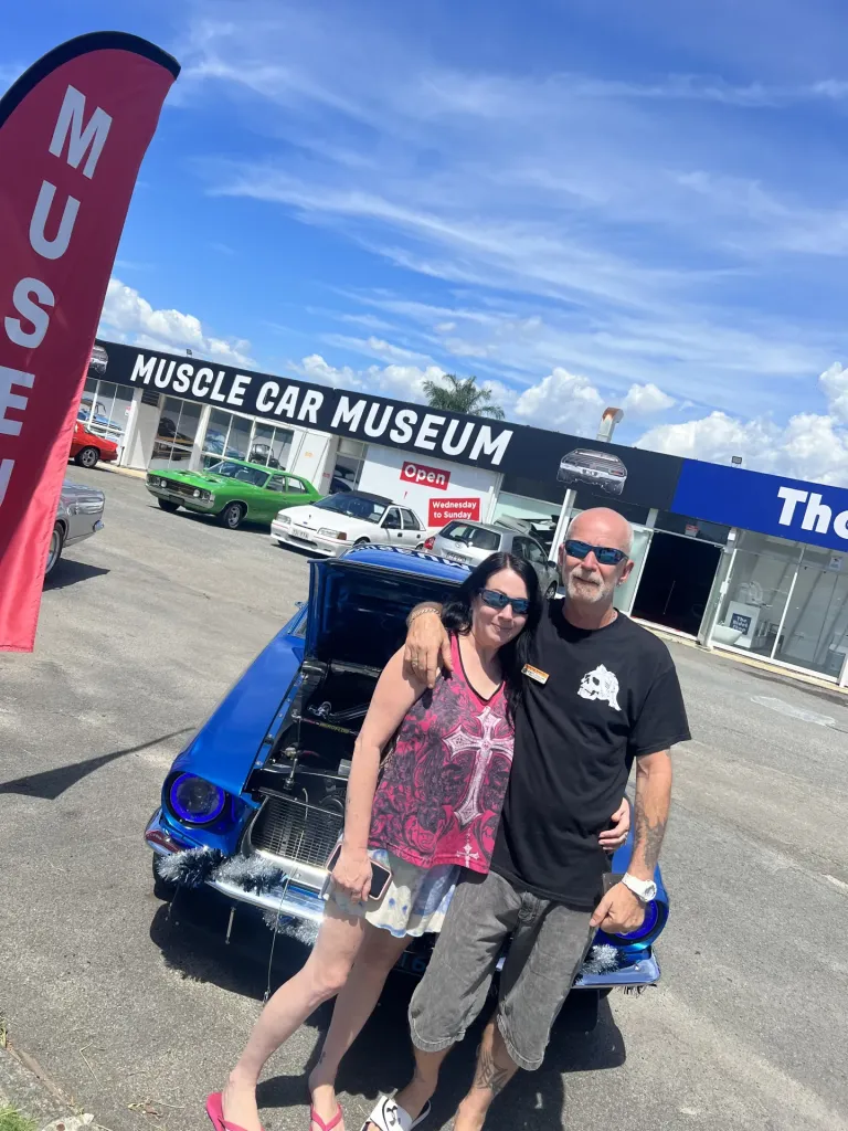 Couple posing in front of a blue car with its bonnet open in the museum car park