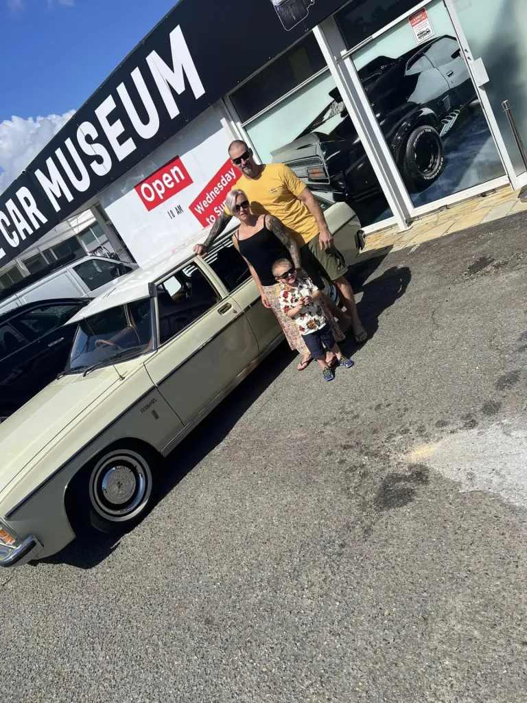Family with a young child standing beside a pale green classic sedan outside the Muscle Car Museum