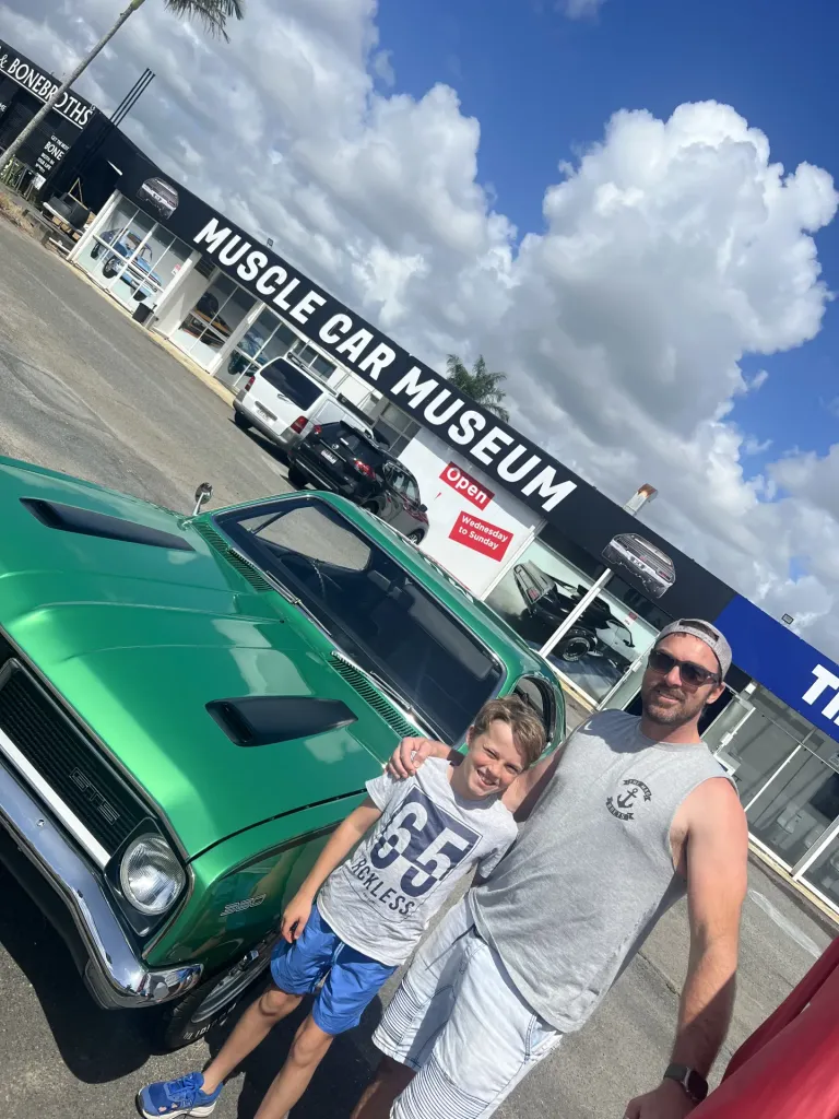 Man and boy posing beside a green classic coupe with bonnet scoops outside the Muscle Car Museum