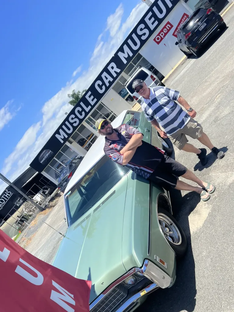Two men leaning on a pale green classic sedan in front of the Muscle Car Museum sign
