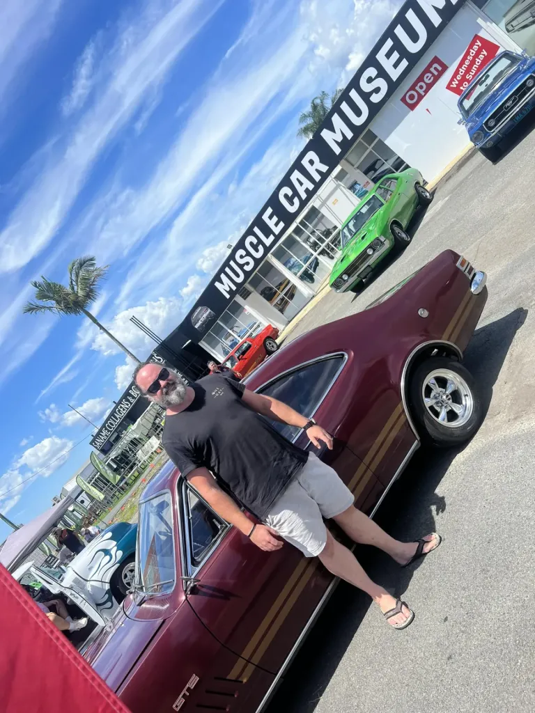 Bearded man leaning on a maroon Holden Monaro GTS coupe with gold stripes outside the Muscle Car Museum