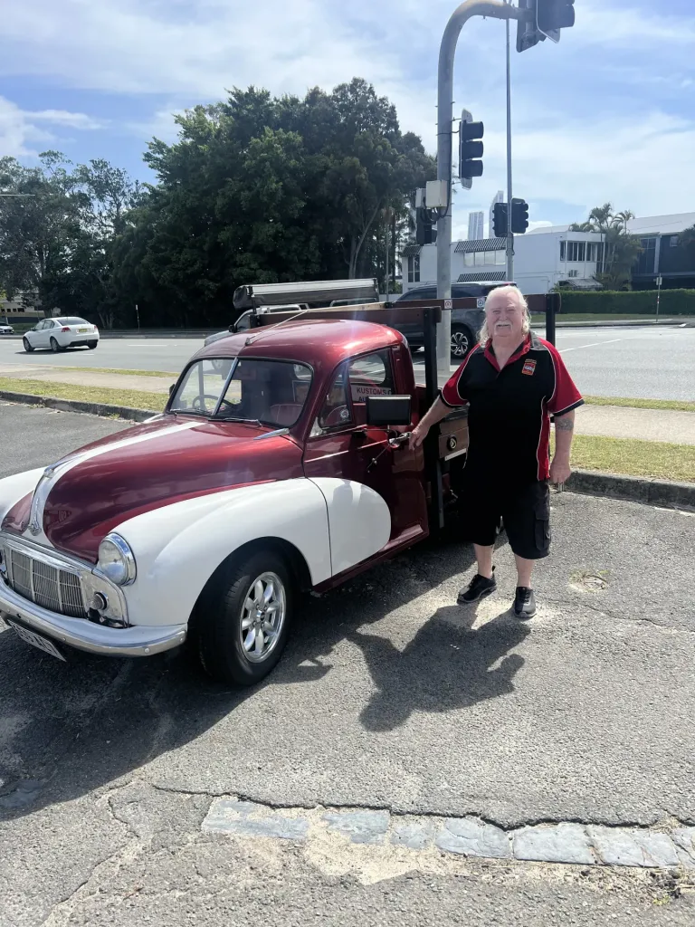 Man standing beside a maroon and white vintage ute parked by a road with traffic lights