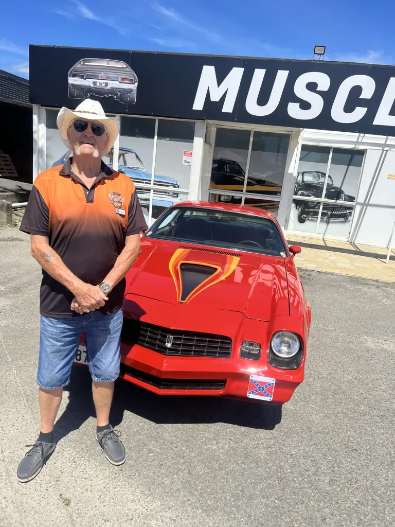 Man in a cowboy hat standing beside a red coupe with a flame-painted bonnet outside the museum entrance