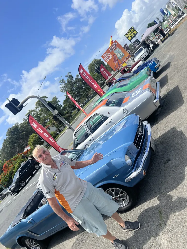 Smiling man gesturing beside a blue 1960s coupe at the front of a row of classic cars at a car meet