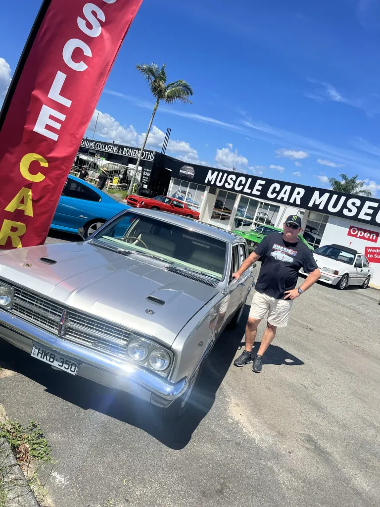 Man posing beside a silver classic Holden sedan in the museum car park, with a red Muscle Car flag banner