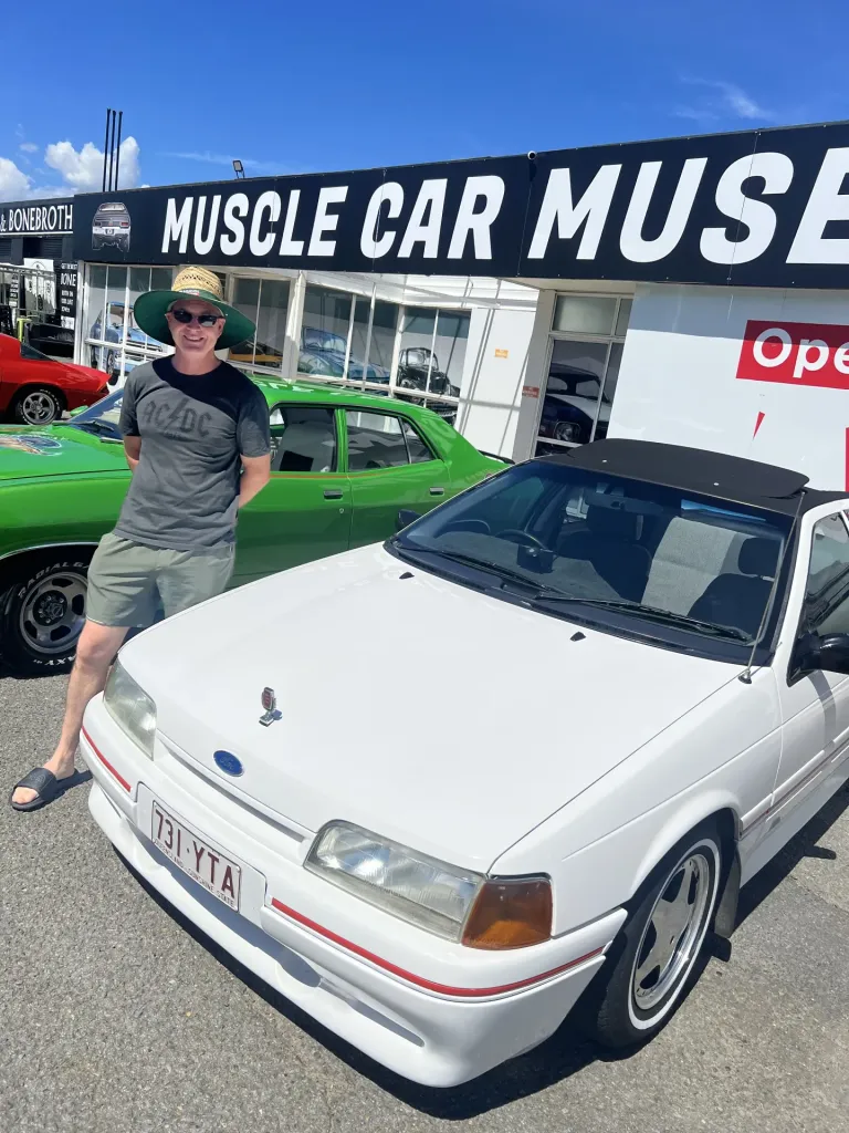 Man in a straw hat beside a white Ford sedan in front of the Muscle Car Museum sign, with a green sedan behind