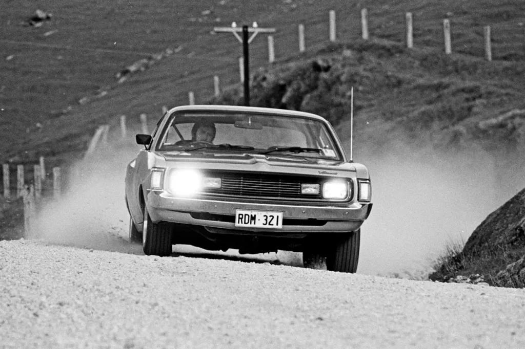 Black and white photo of a Chrysler Valiant Charger driving along a dusty gravel road with headlights on