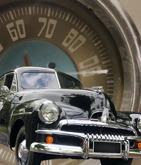 Black Holden FJ sedan with a chrome grille in front of a large vintage speedometer