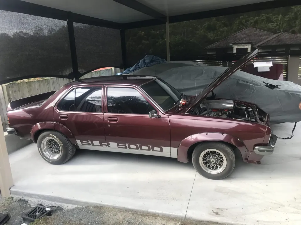 Side view of a maroon Holden Torana SL/R 5000 sedan with its bonnet open under a carport
