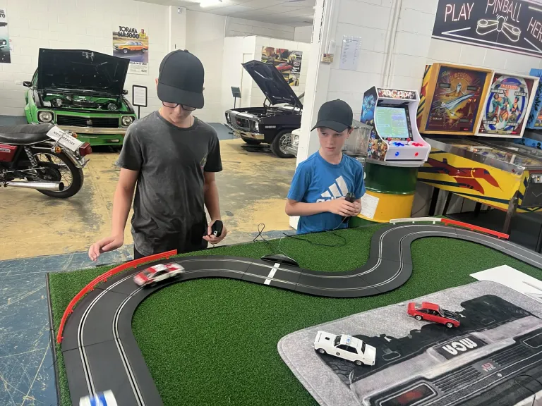 Two boys racing cars on a Scalextric slot car track, pinball machines and classic cars behind