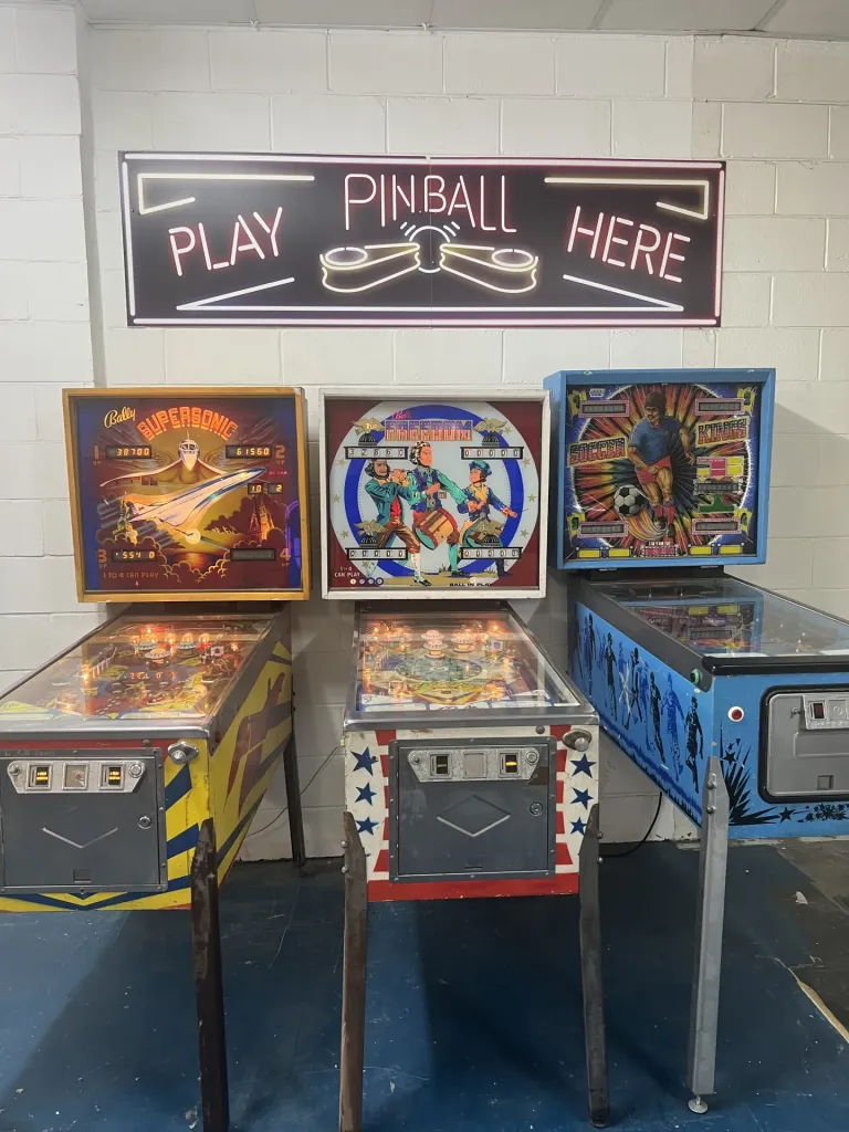 Three vintage pinball machines lined up under a Play Pinball Here neon sign