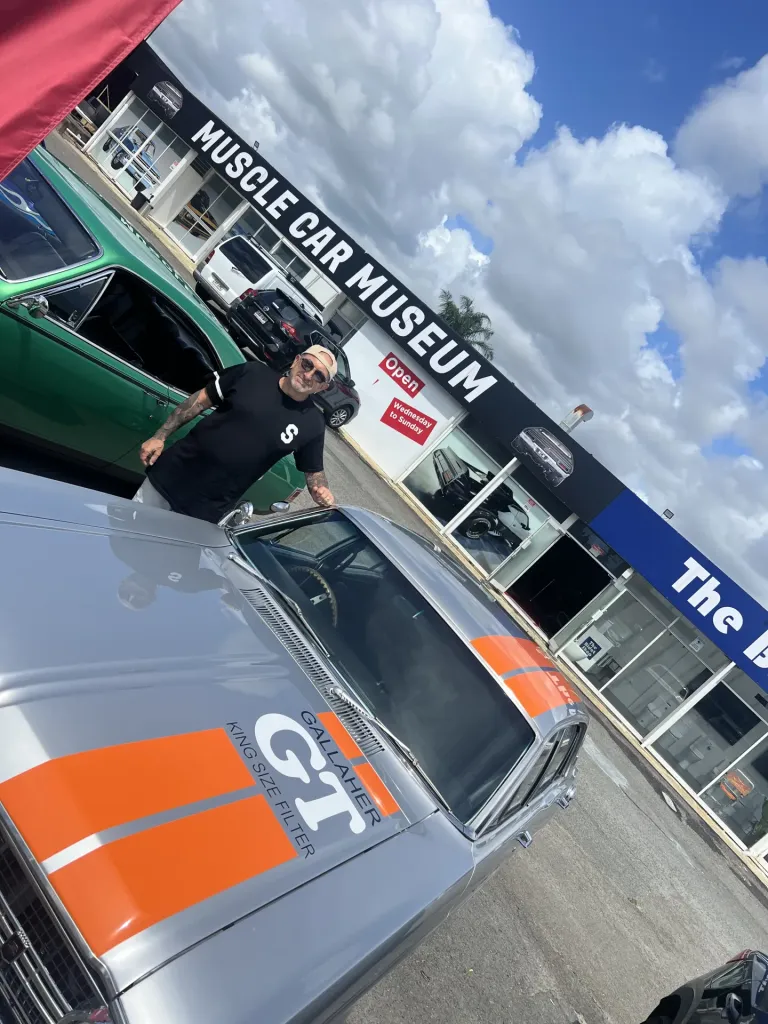 Man leaning on a silver sedan with orange stripes and Gallaher GT livery outside the Muscle Car Museum