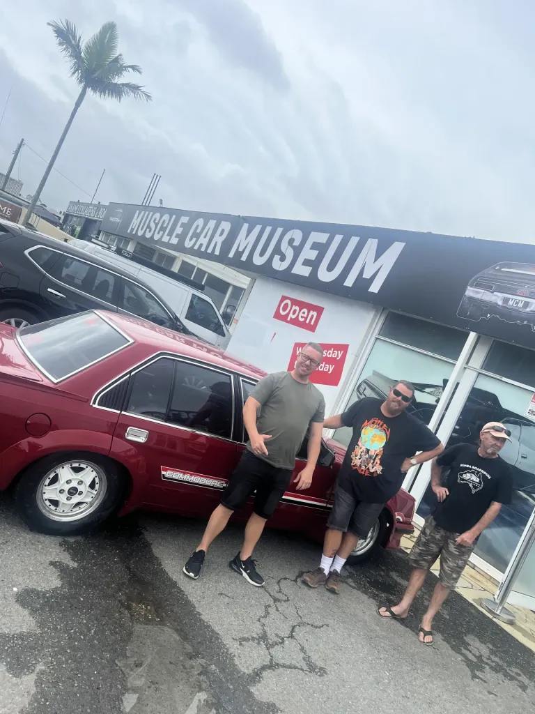 Three men standing beside a red Holden Commodore outside the Muscle Car Museum