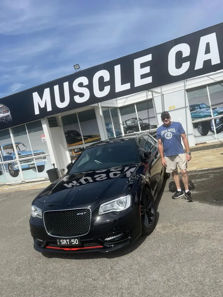 Man in a blue T-shirt standing beside a black Chrysler 300 SRT outside the Muscle Car Museum