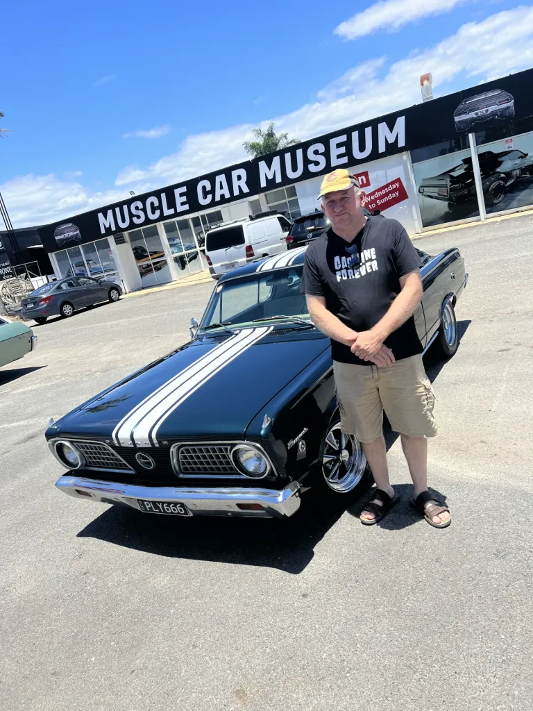 Man in a yellow cap beside a black classic fastback with white racing stripes outside the museum