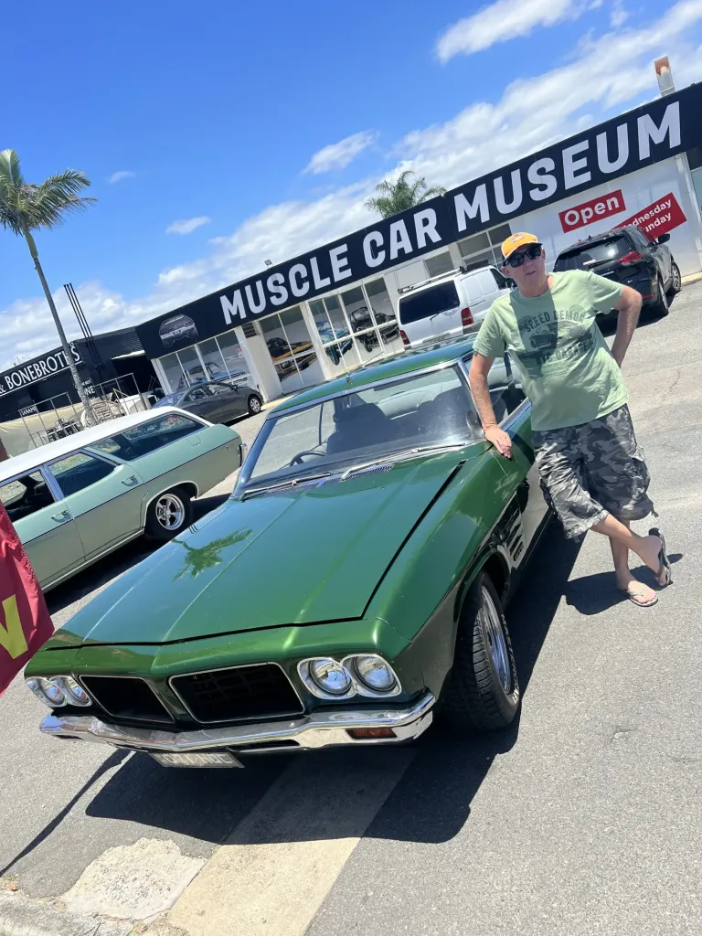 Man in sunglasses leaning on a dark green classic coupe outside the Muscle Car Museum