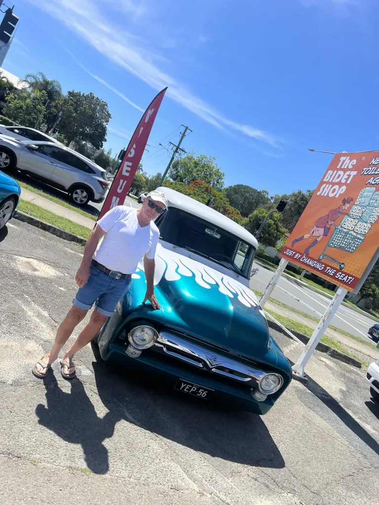 Man in a white cap standing beside a teal 1950s pickup truck with white flame paint in the car park