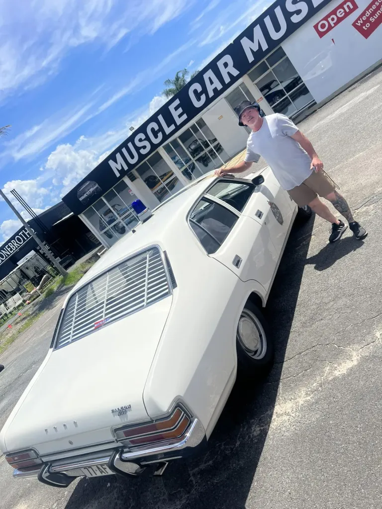 Man in a bucket hat beside a white Ford Falcon sedan with rear window louvres outside the museum